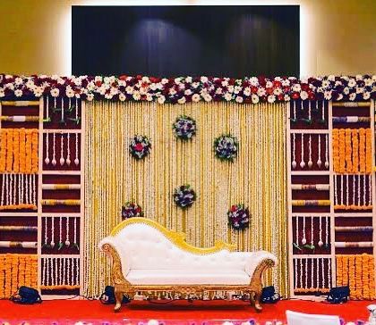 An elaborate stage decoration for a traditional ceremony. This setup uses a backdrop made of floral panels and shelving, with marigold garlands and a classic white chaise lounge for seating.