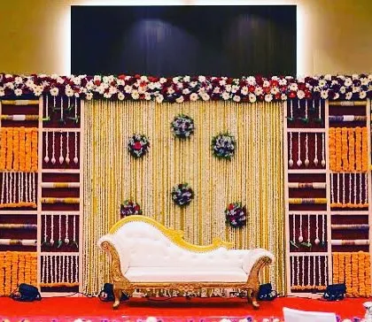 An elaborate stage decoration for a traditional ceremony. This setup uses a backdrop made of floral panels and shelving, with marigold garlands and a classic white chaise lounge for seating.