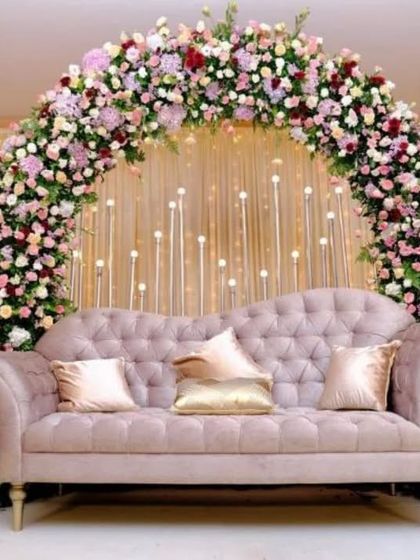 A classic and romantic stage backdrop featuring a lush arch of pink, white, and red roses over a tufted sofa. The background is lit with candle-like bulbs, adding to the warm and inviting feel.