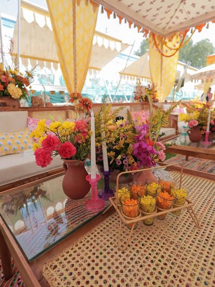 A tablescape from the Rajasthani Haldi, featuring a cane-woven table, colorful florals in a terracotta pot, and a caddy of flower-filled shot glasses for guests.