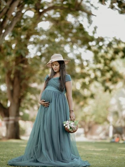 A rustic and charming maternity shoot. The teal off-shoulder gown paired with a straw hat and a basket of flowers creates a lovely, natural look.