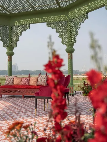 A view from behind the vibrant red flowers lining the aisle, looking towards the couple's seating inside the mandap. This perspective highlights the lushness of the floral design.