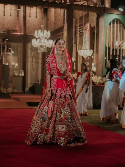 The bride's radiant smile as she walks down the red carpet. The opulent hall with its grand chandeliers provides a fittingly luxurious backdrop for her entrance.