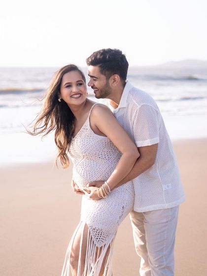 A playful and loving moment between a couple during their beach maternity shoot. The partner hugs the mom-to-be from behind as they share a happy, candid glance.