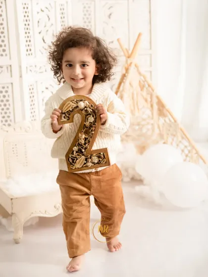 A toddler boy proudly holds up a decorative number "2" to celebrate his second birthday in a neutral, boho-themed studio set.