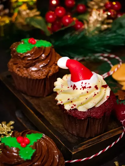 A pair of festive Christmas cupcakes, one with a Santa hat and the other with a holly garnish. The dark, moody lighting creates a cozy and intimate holiday feel.