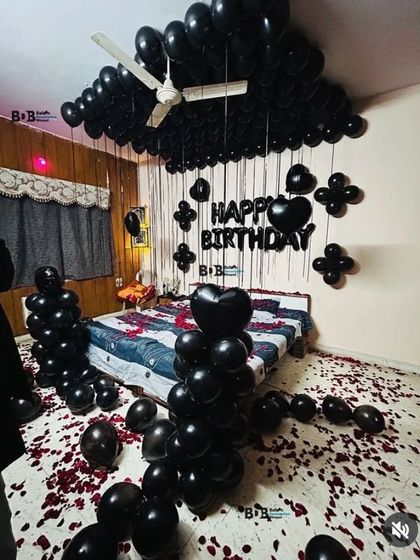 A dramatic all-black balloon decoration for a bedroom, including balloons on the ceiling, heart-shaped balloons, and rose petals on the bed.