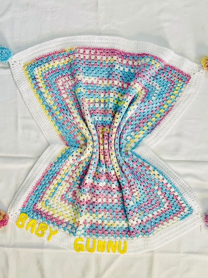 The soft folds of the "Baby Gunnu" blanket highlight the beautiful pastel colors and the texture of the crochet stitches. The personalized name is clearly visible at the top.