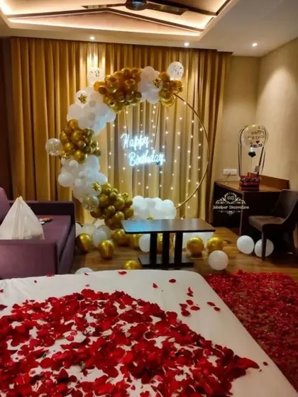 A luxurious hotel room birthday surprise. A gold and white balloon arch with a neon sign is set up by the bed, which is covered in red rose petals for a touch of romance.
