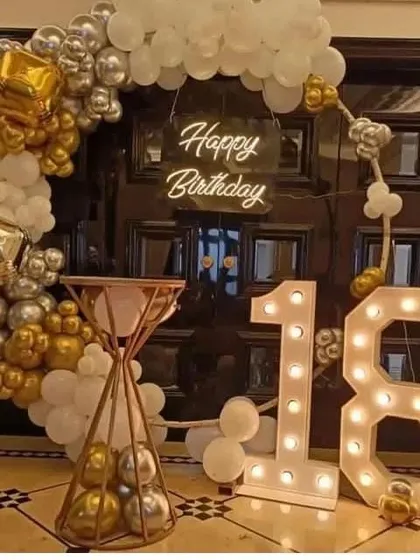 A glamorous 18th birthday entrance decor. This setup features a gold, silver, and white balloon arch over a doorway, a 'Happy Birthday' neon sign, and a large '18' marquee light.