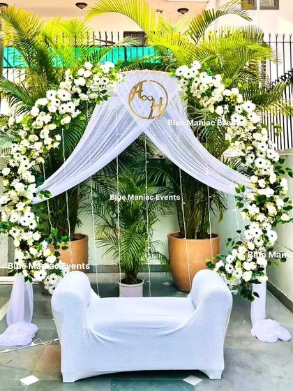 A stunning outdoor day-time setup for a Roka or Nikkah. The circular arch is adorned with white flowers and drapes, beautifully complemented by the natural greenery in the background.