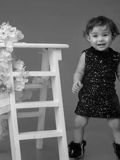 A classic black and white portrait of a toddler in a sparkly dress. The simple composition with the white ladder prop creates a stylish and timeless feel.