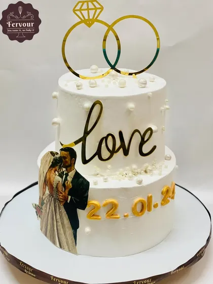 A classic two-tier wedding cake customized with the couple's photo and their special date. The simple white design is accented with edible pearls and a gold ring topper, creating a clean and personal look.