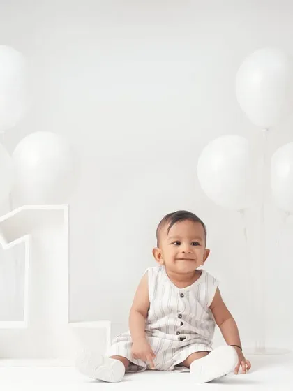 A simple and beautiful first birthday portrait of a baby boy in a minimalist all-white studio with white balloons and a large '1' prop.