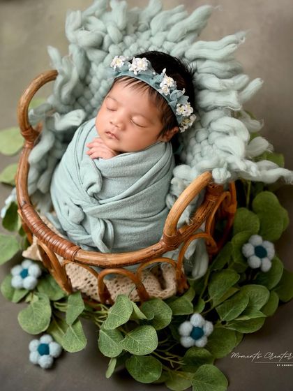 A sleeping beauty in soft blue. This newborn girl, wearing a delicate floral headband, is swaddled and sleeping peacefully in a rustic basket.
