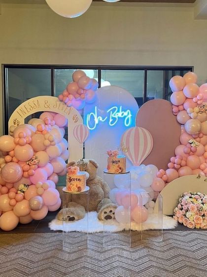 An adorable 'Oh Baby' setup for a twin's baby shower. The decoration features two cakes on clear stands, a large teddy bear, and an abundance of pink balloons.