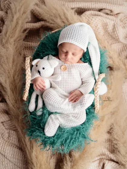 A sleeping newborn in a cozy, textured white romper and matching hat, holding a small knitted toy. This boho-inspired shot uses natural textures like pampas grass for a warm, earthy feel.
