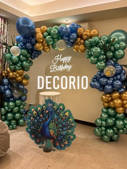 A full view of the peacock-themed birthday decoration, showcasing the complete balloon arch in metallic blues, greens, and golds, along with the peacock cutout and neon sign.
