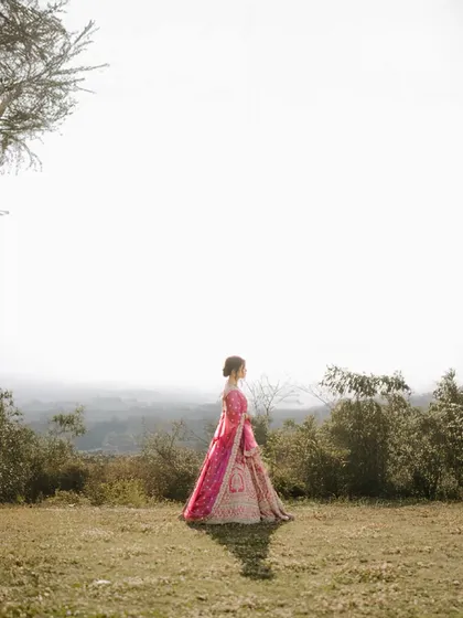 A wide shot of a bride in a vibrant pink lehenga, standing alone in a misty mountain landscape, creating a dreamy and ethereal portrait.