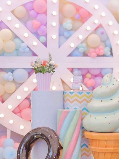 A close-up of the sweet treats corner at a pastel carnival party. Giant props of an ice cream cone, a donut, and a marshmallow twist are set against a lit Ferris wheel backdrop filled with balloons.