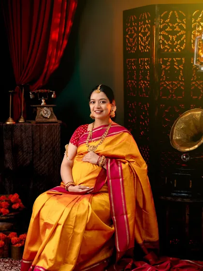 A classic vintage-themed maternity portrait. The mom-to-be is seated gracefully in a yellow and pink silk saree, surrounded by antique props like a gramophone and a classic telephone.