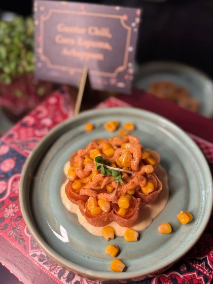 A gourmet take on a simple ingredient: Guntur Chili Corn Lagsana with Achappam. This dish combines the heat of Guntur chili with the sweetness of corn, served on a crispy, flower-shaped achappam for a delightful texture.