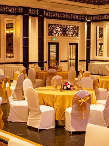 A view of the guest seating in a grand ballroom. The tables are dressed in gold linens with white chair covers and gold sashes, creating a classic and opulent look for a wedding reception.