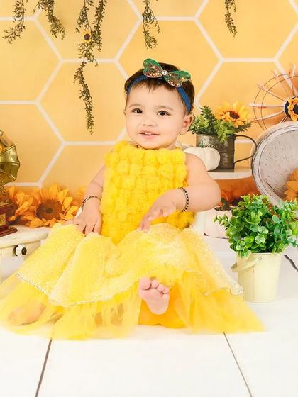 A baby girl in a fluffy yellow dress sits among sunflowers, looking like a little ray of sunshine.
