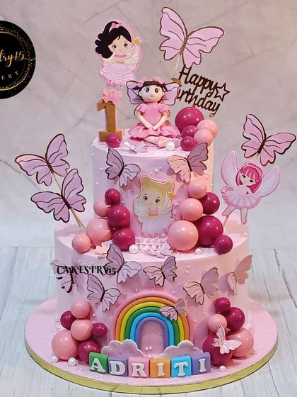 A full view of the two-tier pineapple cake for a first birthday. The design is a fantasy of pink, with a fondant girl, butterflies, and a rainbow, creating a magical and sweet centerpiece.