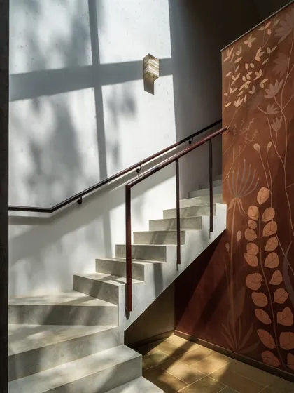 Sunlight casts dramatic shadows across a concrete staircase, highlighting a beautiful, nature-inspired mural on the wall. The interplay of light, texture, and art creates a visually stunning architectural detail.