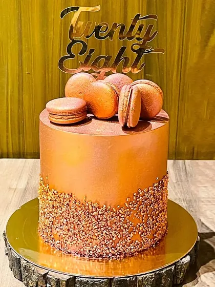 An elegant and modern 28th birthday cake. This custom chocolate walnut cake has a sleek metallic bronze finish, crunchy praline at the base, and is topped with delicate, handmade macarons.