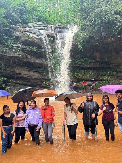 The group enjoying the rain at a waterfall, with colorful umbrellas adding to the cheerful monsoon atmosphere.