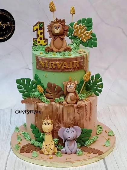 A wild two-tier jungle-themed cake for a first birthday. This cake is adorned with adorable fondant animals including a lion, monkey, giraffe, and elephant amidst lush green leaves.