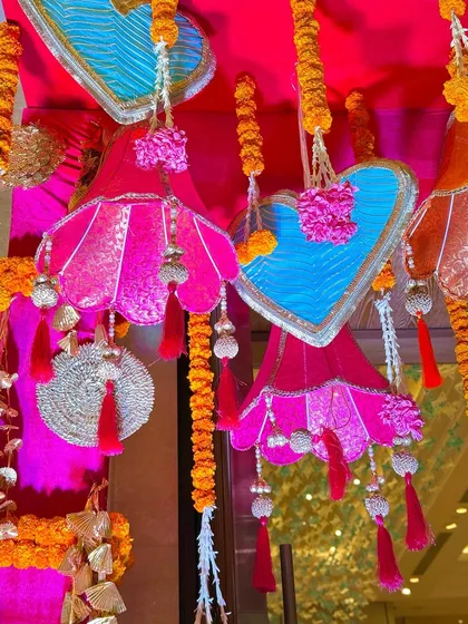 Intricate hanging decorations designed for a Garba night. This image shows custom-made fabric lampshades and heart-shaped ornaments in bright pink and blue, accented with marigold garlands and silver tassels.