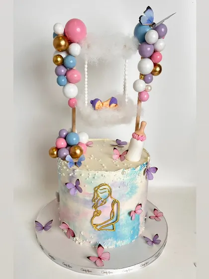 A whimsical baby shower cake with a watercolor pastel finish. The cake is adorned with butterflies and features a unique topper of a baby sleeping in a swing suspended between two balloon columns.