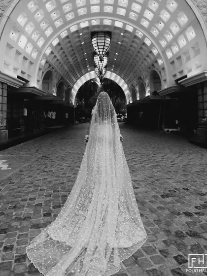 A portrait of timeless elegance. We work with you and your photography team to stage moments that feel like art, like this bride's stunning portrait featuring a dramatic, floor-length veil in a grand architectural hallway, captured in classic black and white.