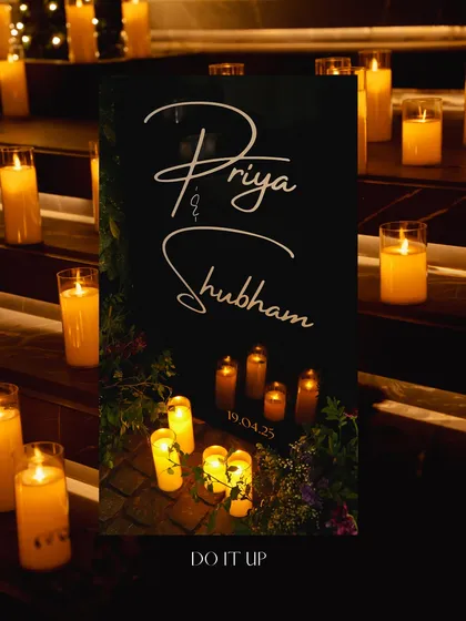 The welcome sign for the Sangeet night in Rishikesh. The couple's names are elegantly scripted on a black sign, illuminated by the soft, warm glow of surrounding candles.