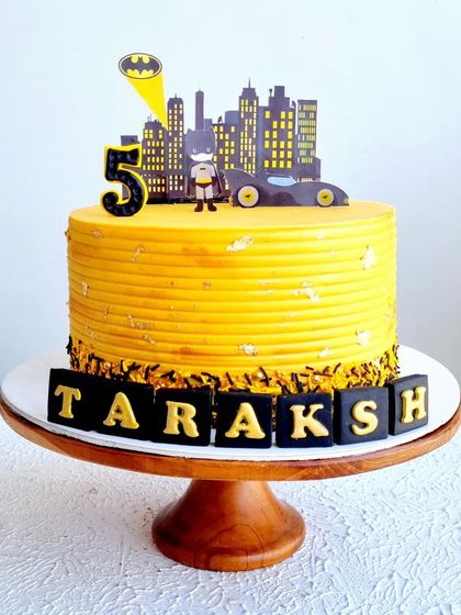 A Batman-themed cake with a yellow buttercream texture and a city skyline topper. The name is spelled out in fondant blocks.