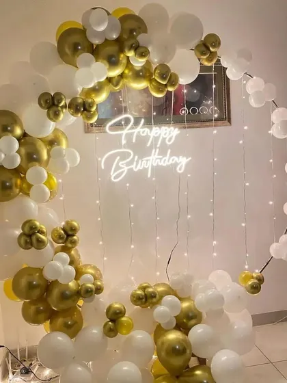 This modern and chic birthday backdrop features a 'Happy Birthday' neon sign inside a circular frame. The frame is asymmetrically decorated with metallic gold and white balloons, with fairy lights adding a warm glow.