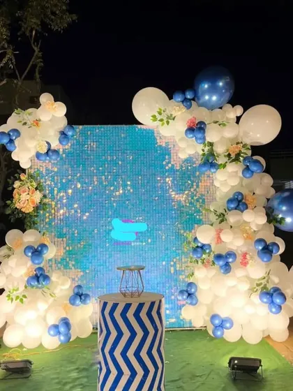 An outdoor party backdrop with a blue shimmer wall and a white balloon garland.