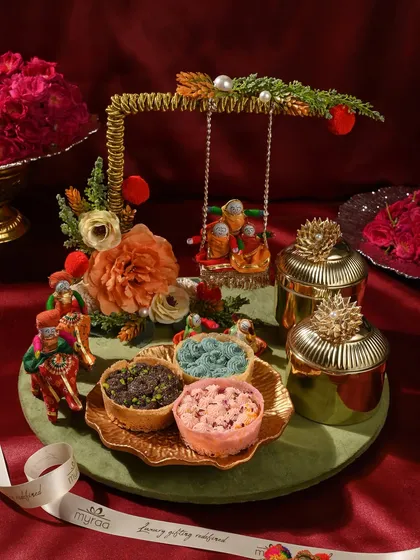 This whimsical Teej hamper features a miniature swing (jhoola), colourful ghewars, and ornate golden jars. It’s a playful and traditional gift that captures the essence of the festival.