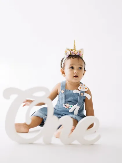 Celebrating year one with a touch of magic. This little one, wearing a unicorn headband and cute denim overalls, sits proudly next to a "One" sign, marking her very first birthday.