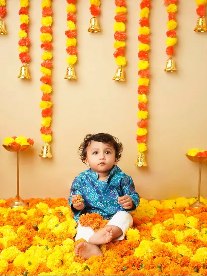A vibrant festive photoshoot of a baby boy dressed in a traditional blue kurta, sitting in a sea of bright orange and yellow marigold flowers, with festive decorations hanging in the background.