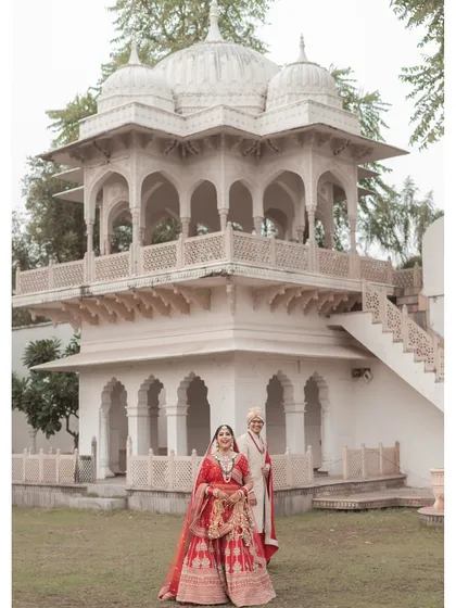 A classic portrait of the bride and groom standing before a beautiful white marble gazebo. The bride's red lehenga provides a stunning contrast, creating a timeless image of their royal wedding day.