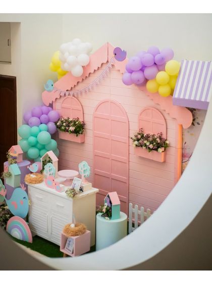 A whimsical birdhouse-themed party setup, featuring a large pink house, bird cutouts, and nests, all in a soft pastel palette.