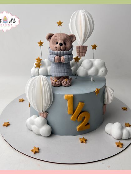 Halfway to one! A dreamy half-birthday cake featuring a cozy teddy bear, hot air balloons, and fluffy clouds in a soft blue and grey palette.