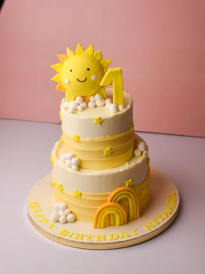 A burst of sunshine for a first birthday celebration. This cheerful two-tier cake features a bright yellow and white design with fondant suns, clouds, and rainbows, perfect for celebrating your little one's big day.