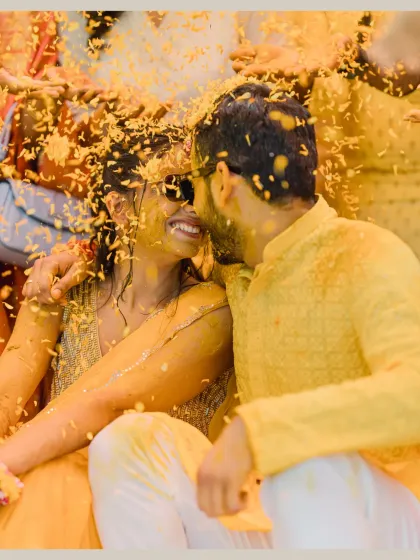 An intimate moment amidst the Haldi chaos. The couple, covered in turmeric and flowers, share a quiet, loving glance.