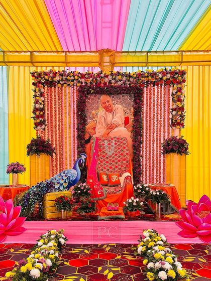 An elaborate indoor setup for a Guruji satsang, featuring a canopy of colorful drapes. The main seating area is richly decorated with floral garlands and traditional elements.