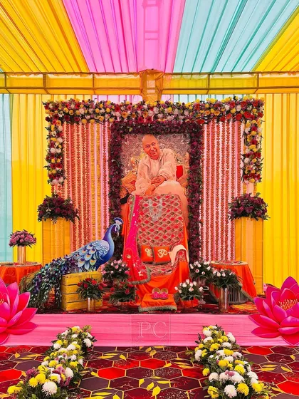 An elaborate indoor setup for a Guruji satsang, featuring a canopy of colorful drapes. The main seating area is richly decorated with floral garlands and traditional elements.